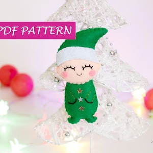 May include: PDF pattern for a felt Christmas elf ornament. The elf is wearing a green hat and a green and white outfit with gold stars. The elf has a smiling face with closed eyes.