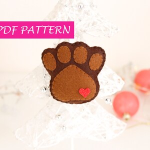 May include: A brown felt paw print ornament with a red felt heart in the center. The ornament is hanging on a white, lacy Christmas tree.