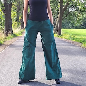 May include: A woman wearing a black tank top and dark green wide-leg pants with cargo pockets. She is standing on a paved road with trees on either side.