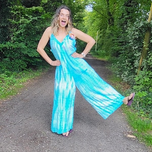 May include: A turquoise tie-dye jumpsuit with spaghetti straps. The wide-leg jumpsuit features a V-neck and a colorful patch on the chest. The model is wearing sandals and standing on a paved path surrounded by trees.