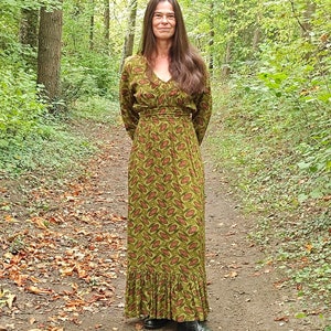 May include: A woman wearing a long, green maxi dress with a red and green leaf pattern. The dress has a V-neckline and long sleeves. She is standing in a woodland setting with fallen leaves on the ground.