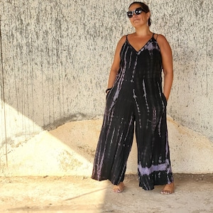 May include: A person wearing a black and purple tie-dye jumpsuit with a V-neck and thin straps. They are wearing sunglasses and standing in front of a textured wall.