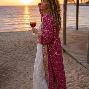 May include: A person wearing a long, fuchsia embroidered robe with gold detailing, holding a glass of red wine on a beach at sunset. They are also wearing a white dress. The scene is set on a sandy beach with wooden structures.