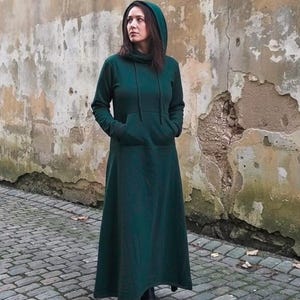 May include: A dark green hooded dress with a long skirt. The dress has a kangaroo pocket and a hood. The person is wearing black boots. The background is a weathered wall and a brick walkway.