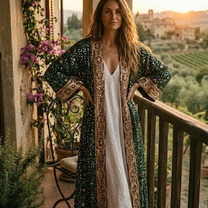 May include: A long, dark green sequined kimono with gold trim is worn over a white dress. The kimono has wide sleeves and a detailed border. The background shows a balcony with a view of a landscape and a sunset.