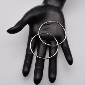 May include: Two silver hoop earrings. The earrings are circular and have a shiny, metallic finish. They are displayed on a black hand against a light gray background. The hoops are a classic jewelry design.