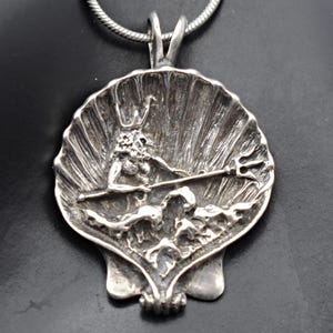 May include: A silver pendant featuring a detailed depiction of a mythological figure, possibly Poseidon or Neptune, holding a trident within a seashell design. The pendant is suspended from a silver chain, showcasing intricate craftsmanship and a nautical theme.