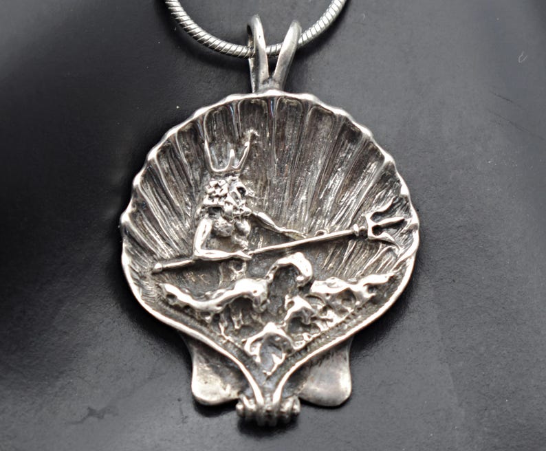 May include: A silver pendant featuring a detailed depiction of a mythological figure, possibly Poseidon or Neptune, holding a trident within a seashell design. The pendant is suspended from a silver chain, showcasing intricate craftsmanship and a nautical theme.