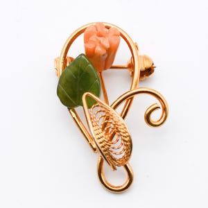 May include: A gold-toned brooch featuring a floral design. The brooch includes a carved coral-colored flower, a green leaf, and a woven gold-colored element. The design is set against a white background.