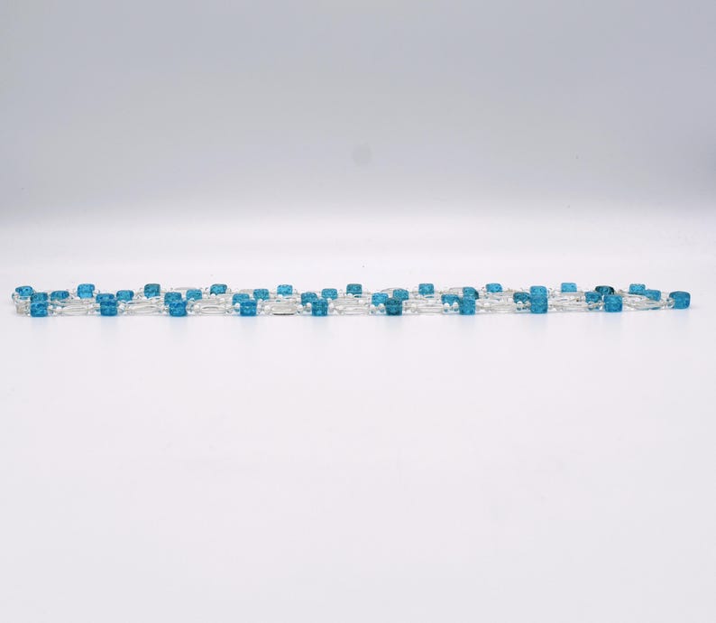 Op de afbeelding: Een zilveren ketting met afwisselend heldere en blauwe vierkante kralen.