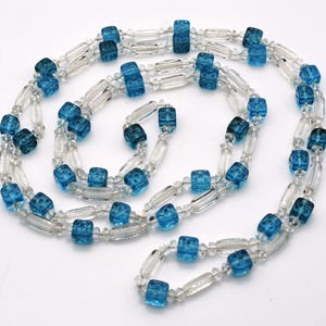 Op de afbeelding: Een lange ketting gemaakt van heldere en blauwe glazen kralen. De blauwe kralen zijn kubusvormig en hebben een getextureerd oppervlak. De ketting is ongeveer 76 cm lang.