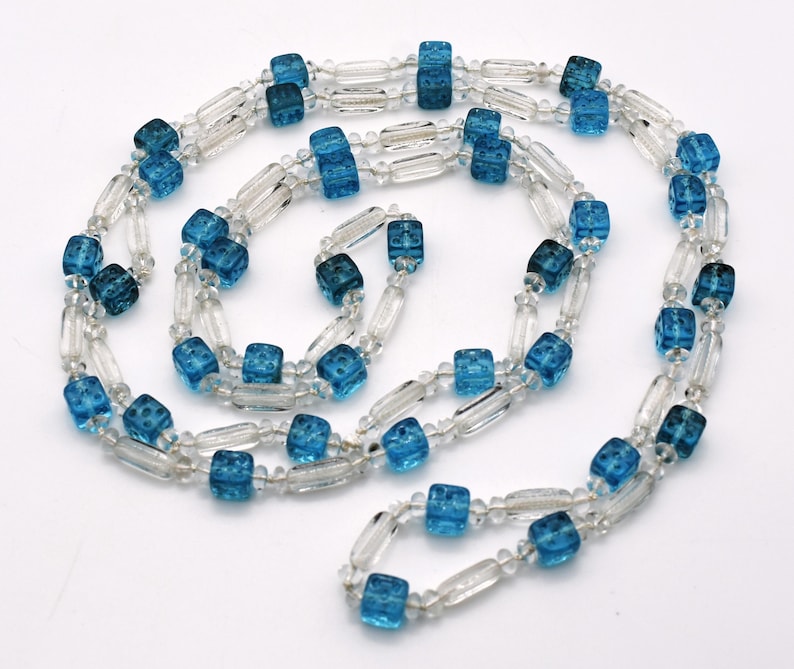 Op de afbeelding: Een lange ketting gemaakt van heldere en blauwe glazen kralen. De blauwe kralen zijn kubusvormig en hebben een getextureerd oppervlak. De ketting is ongeveer 76 cm lang.