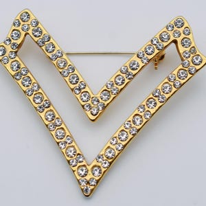 Peut inclure: Broche dorée en forme de double chevron, incrustée de strass transparents. L'épingle a un style classique et élégant, idéale pour ajouter de l'éclat à n'importe quelle tenue. La broche mesure environ 5 cm de large.