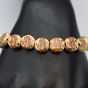 May include: A brown bracelet with beige beads featuring a floral design. Each bead has a brown flower with green leaves.