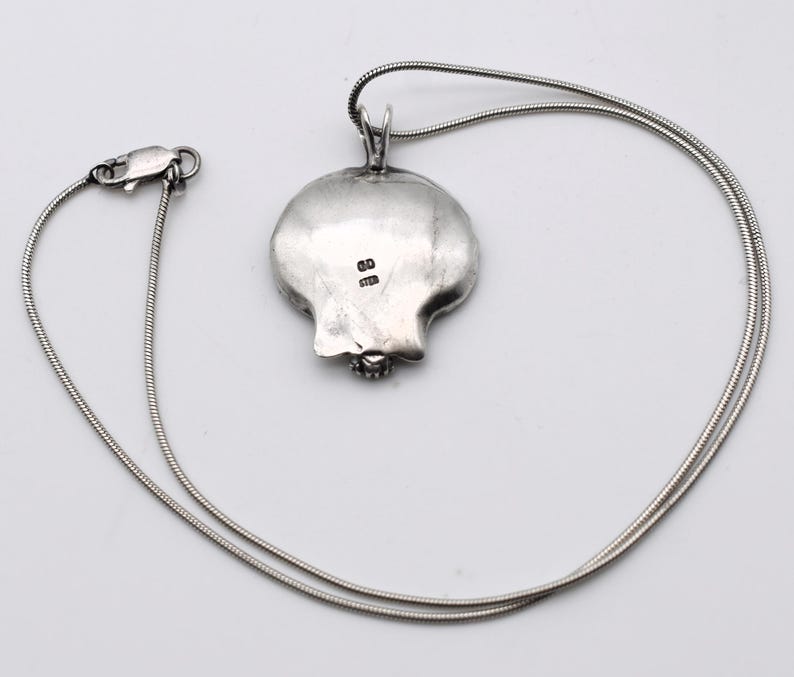 May include: A silver pendant necklace featuring a pomegranate-shaped pendant. The pendant is detailed with a textured surface and a small gemstone at the base. The necklace has a delicate, snake chain and a secure clasp.