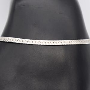 Könnte beinhalten: Ein silberfarbenes Fischgrätenarmband. Das Armband ist dünn und flexibel, mit einem flachen, gewebten Design. Das Metall hat eine helle, reflektierende Oberfläche. Das Armband wird vor einem dunklen Hintergrund präsentiert.