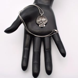 May include: A silver-toned necklace featuring a seashell pendant. The pendant has a detailed design with a textured surface. The necklace is displayed on a black hand against a white background. The chain is delicate and the clasp is visible.