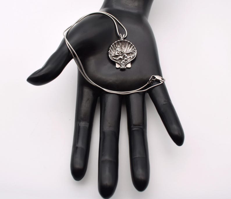 May include: A silver-toned necklace featuring a seashell pendant. The pendant has a detailed design with a textured surface. The necklace is displayed on a black hand against a white background. The chain is delicate and the clasp is visible.