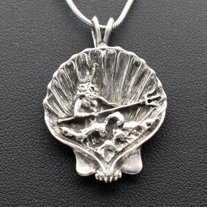 May include: A silver pendant featuring a detailed depiction of a mythological figure, possibly Poseidon or Neptune, holding a trident within a seashell design. The pendant hangs from a silver chain. The pendant is approximately 1.5 inches in diameter.