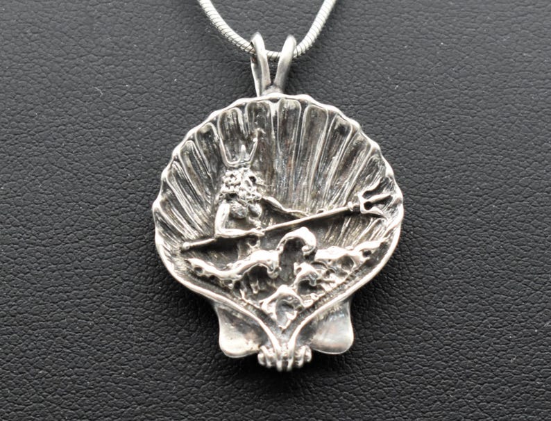 May include: A silver pendant featuring a detailed depiction of a mythological figure, possibly Poseidon or Neptune, holding a trident within a seashell design. The pendant hangs from a silver chain. The pendant is approximately 1.5 inches in diameter.