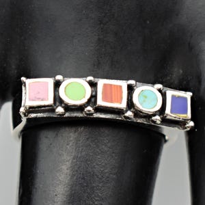 May include: A silver ring with a row of colorful inlaid stones. The stones are square and round, in shades of pink, green, orange, turquoise, and blue. The ring is on a black background.