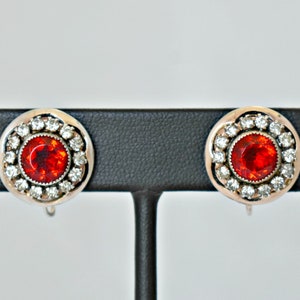 May include: A pair of silver clip-on earrings with a red rhinestone surrounded by a halo of smaller clear rhinestones.