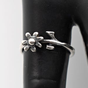May include: A silver ring featuring a daisy flower design with a central bead and a branch-like band. The ring is displayed on a black surface, showcasing its intricate details and floral theme. The ring's design is delicate and nature-inspired.