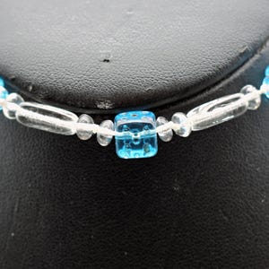 Op de afbeelding: Een ketting van blauwe en heldere glazen kralen met vierkante kralen. De ketting ligt op een zwart oppervlak.