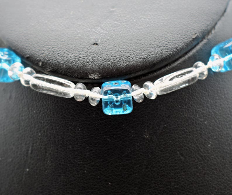 Op de afbeelding: Een ketting van blauwe en heldere glazen kralen met vierkante kralen. De ketting ligt op een zwart oppervlak.