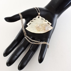 May include: A silver chain necklace with a large, white, carved shell pendant. The pendant features a floral design and two small pearls.