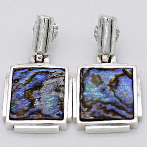 Peut inclure: Paire de boucles d'oreilles argentées avec incrustations rectangulaires en coquillage d'ormeau. Les coquillages présentent des reflets irisés bleus, verts et bruns. Fermeture à tige et design géométrique. Mesurent environ 2,5 cm de long.