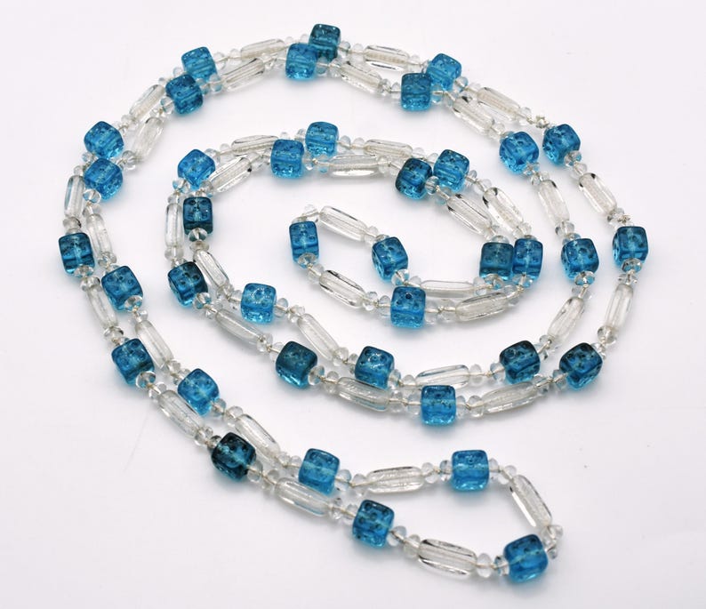 Op de afbeelding: Een lange ketting gemaakt van heldere en blauwe glazen kralen. De kralen zijn vierkant en rechthoekig van vorm.