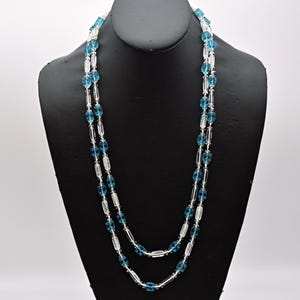 Op de afbeelding: Een dubbele ketting met heldere en blauwe glazen kralen. De kralen zijn ovaalvormig en hebben een facetgeslepen ontwerp.
