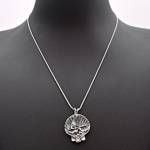 May include: A silver-toned pendant necklace featuring a detailed seashell design. The pendant showcases a sculpted scene within the shell, suspended from a delicate silver chain. The necklace is displayed on a black mannequin.
