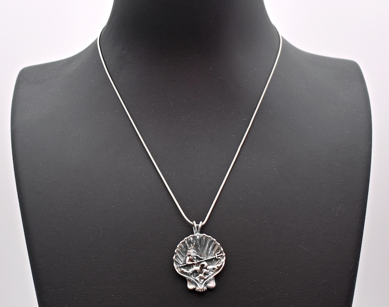 May include: A silver-toned pendant necklace featuring a detailed seashell design. The pendant showcases a sculpted scene within the shell, suspended from a delicate silver chain. The necklace is displayed on a black mannequin.