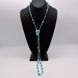 Op de afbeelding: Een lange, gelaagde ketting met blauwe en heldere glazen kralen. De ketting is in het midden geknoopt en hangt tot aan de borst.