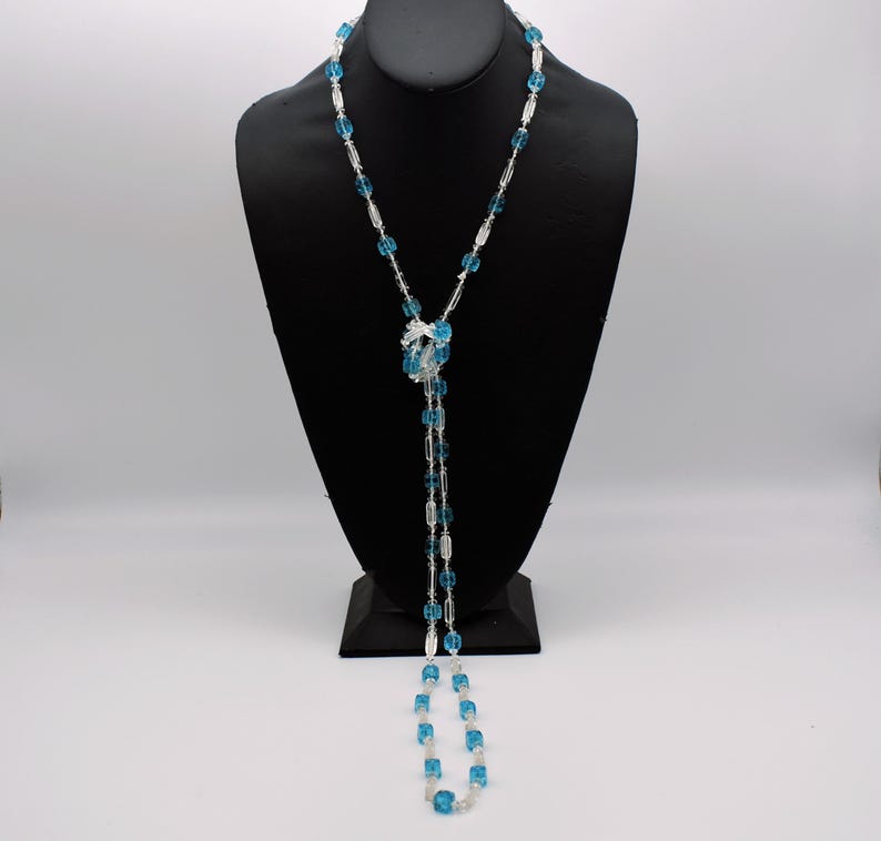 Op de afbeelding: Een lange, gelaagde ketting met blauwe en heldere glazen kralen. De ketting is in het midden geknoopt en hangt tot aan de borst.