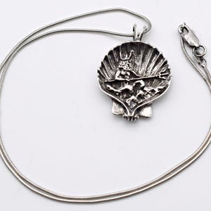 May include: A silver-toned pendant necklace featuring a detailed seashell design. The pendant showcases a sculpted figure with a trident, set within the shell's interior. The necklace has a delicate, snake chain and a secure clasp.