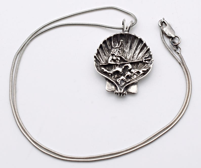May include: A silver-toned pendant necklace featuring a detailed seashell design. The pendant showcases a sculpted figure with a trident, set within the shell's interior. The necklace has a delicate, snake chain and a secure clasp.