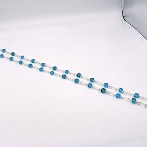 Op de afbeelding: Een lange ketting met afwisselend blauwe en heldere kralen. De kralen zijn gefacetteerd en hebben een vierkante vorm.