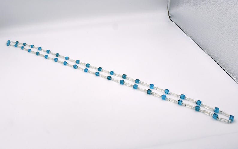 Op de afbeelding: Een lange ketting met afwisselend blauwe en heldere kralen. De kralen zijn gefacetteerd en hebben een vierkante vorm.