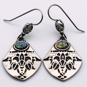 May include: A pair of silver-toned earrings. Each earring features a diamond-shaped pendant with a cut-out floral design and a small, oval-shaped, colorful stone. The earrings have a hook closure.