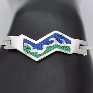 May include: A silver bracelet featuring a central chevron-shaped link with inlaid crushed stone in blue and green. The design depicts stylized wave patterns separated by silver lines, with a polished metallic finish on the surrounding band.