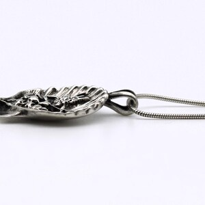 May include: A silver pendant necklace featuring a detailed, leaf-shaped pendant with floral and textured elements. The pendant is attached to a thin, silver chain. The design is intricate and the metal has a polished finish.