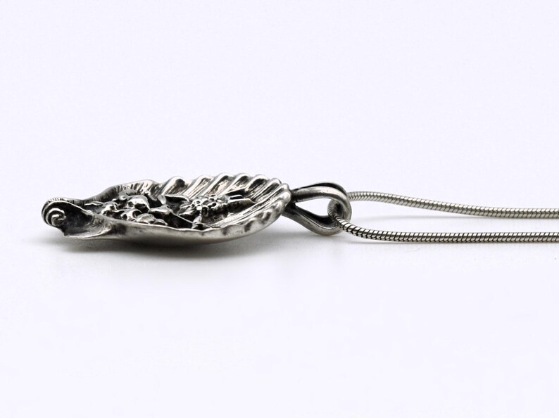 May include: A silver pendant necklace featuring a detailed, leaf-shaped pendant with floral and textured elements. The pendant is attached to a thin, silver chain. The design is intricate and the metal has a polished finish.
