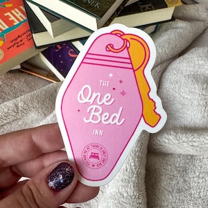 Puede incluir: Pegatina en forma de llave rosa y amarilla con el texto "The One Bed Inn" y una peque&ntilde;a ilustraci&oacute;n de una cama con un coraz&oacute;n. La pegatina est&aacute; en una mano con esmalte de u&ntilde;as de purpurina.