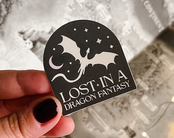 May include: A black and white sticker with a white dragon flying in front of a crescent moon and stars. The sticker says "Lost in a Dragon Fantasy".