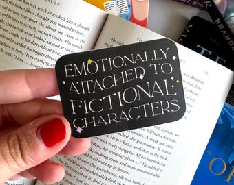 Puede incluir: Una pegatina rectangular negra con esquinas redondeadas que presenta el texto blanco "EMOTIONALLY ATTACHED TO FICTIONAL CHARACTERS" y pequeños acentos de estrellas coloridas, sostenida sobre un libro abierto rodeado de otras portadas.