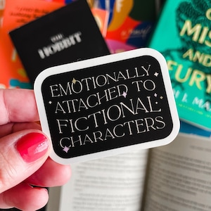 May include: A black and white sticker with the text "EMOTIONALLY ATTACHED TO FICTIONAL CHARACTERS" is held in front of a stack of books. The sticker has a white border and star accents. Books with colorful covers are visible.
