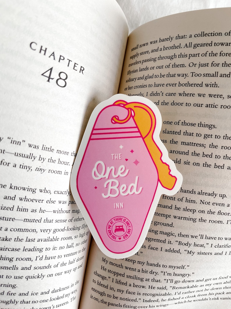 Puede incluir: Una pegatina en forma de llave rosa y amarilla con el texto "The One Bed Inn" y una peque&ntilde;a ilustraci&oacute;n de una cama. La pegatina est&aacute; colocada en una p&aacute;gina de un libro con el texto "Cap&iacute;tulo 48" visible.